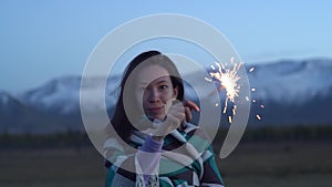 An Asian girl dressed in a blanket holds a sparkler in her hands. Sunset, snowy mountains in the background