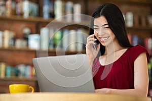 Asian Girl Freelancer Talking On Cellphone While Working On Laptop In Cafe