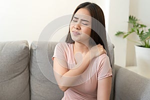 Asian girl having shoulder pain sitting on  sofa at home. young girl has pain from the muscles, Health and illness concepts