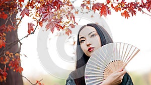 Beautiful young Asian woman with fan on background of red maple