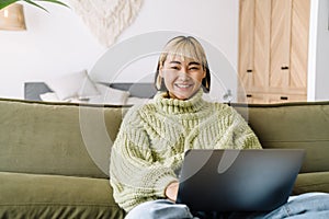 Asian girl laughing and working with laptop while resting on sofa