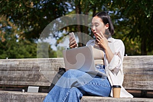 Asian girl looks surprised at smartphone screen, checking mobile phone notifications and looking excited, sitting with