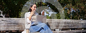 Asian girl looks surprised at smartphone screen, checking mobile phone notifications and looking excited, sitting with