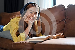 Asian girl lying on sofa, listening to music through headphones and reading a novel