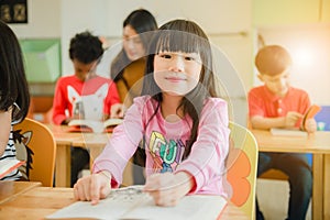 Asian girl reading a book smiling at the camera. Row of multiethnic elementary students reading book in classroom at school.