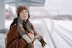 Asian girl in sheepskin coat waiting for train at railway station in snowy weather. Public transport