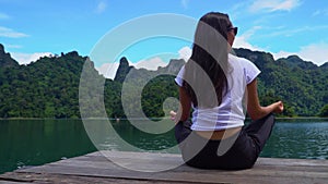 Asian girl sits on a pier in a lotus pose on a lake with a beautiful view.