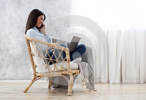 Asian Girl Sitting In Armchair, Working With Laptop And Talking On Cellphone