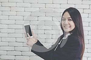 Asian girl smiles and uses her hand to point smartphone, background white brick wall, Online Trading Concepts and Auctions