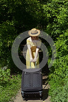 Asian girl in straw hat and yellow dress with big black suitcase among green trees. Summer vacation.