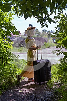 An Asian girl in a straw hat and a yellow dress is carrying a large black suitcase along the road among green trees