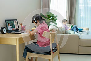 Asian girl student video conference e-learning with teacher and classmates on computer and her sibling reading book insofa in