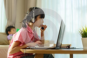 Asian girl student video conference e-learning with teacher and classmates on computer and her sibling reading book in sofa in