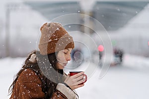 Asian girl traveler drinking hot tea trying to keep warm on city railway station on snowy winter day