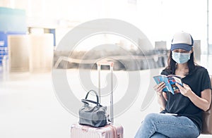 Asian girl traveler wearing surgical mark with carry on luggage in international airport reading a book while waiting for her