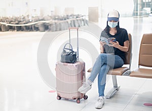 Asian girl traveler wearing surgical mark with carry on luggage in international airport reading a book while waiting for her