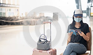 Asian girl traveler wearing surgical mark with carry on luggage in international airport reading a book while waiting for her