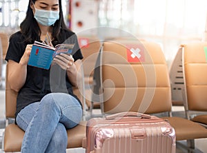 Asian girl traveler wearing surigal mark with carry on luggage in international airport reading a book while waiting for her