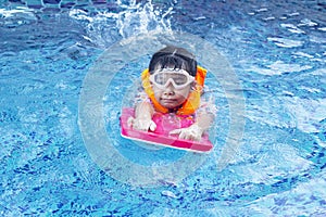 Asian girl wearing an orange life jacket and swimming goggles holding a kickboard is practicing to float happily in the pool.
