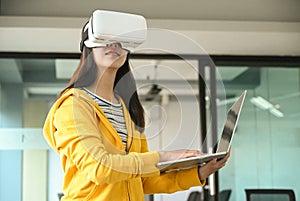 Asian girl wearing a yellow shirt, using VR headset. She is holding laptop and typing a keyboard