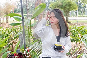 An Asian girl in white coat sitting and holding a black cup of coffee in her hand on the white bench under the morning light.