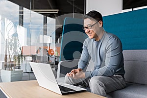 Asian with glasses, working in the office, making a video call with a laptop, smiling and rejoicing, communicating remotely with