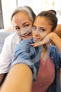 Asian grandmother embracing biracial teenage granddaughter at home, both smiling at camera