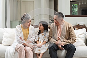 Asian grandparent with little cute grandchild girl sitting on sofa, talking, smiling together. Adorable granddaughter listen to