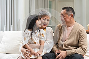 Asian grandparent with little cute grandchild girl sitting on sofa, talking, smiling together. Adorable granddaughter listen to