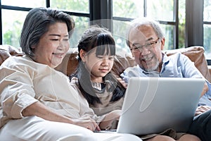 Asian grandparent with little young cute grandchild sitting on sofa playing laptop together. Little granddaughter teach senior eld