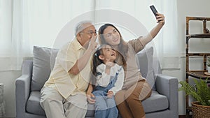 Asian grandparents laughing taking selfie with granddaughter on sofa at home by mobile