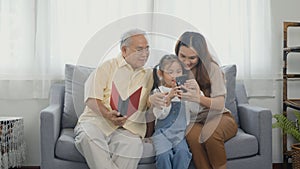 Asian grandparents laughing taking selfie with granddaughter on sofa at home by mobile