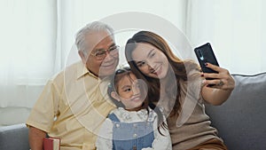 Asian grandparents laughing taking selfie with granddaughter on sofa at home by mobile