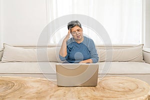 Asian gray hair old woman looking at laptop computer monitor, place her hand atop her head while sit on sofa in the living room