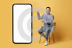 Asian guy pointing at smartphone with white screen while sitting on chair