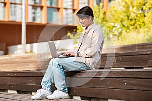 Asian guy sitting on bench at urban area, using laptop