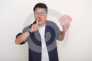 Asian guy standing and holding lots of money gesturing to keep it secret