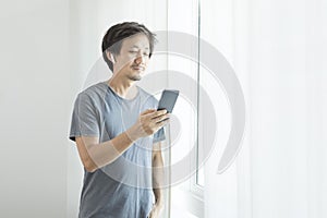 Asian guy using voice assistant on cellphone with earpod by the window.