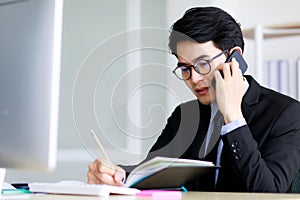 Asian handsome business man wearing eyeglasses, formal black suit with necktie, working and writing on notebook while talking or