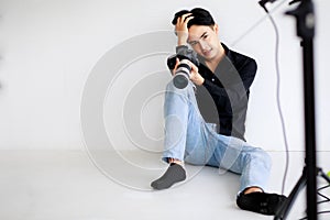 Asian handsome male model wearing casual black shirt with jeans, holding a camera, sitting on white background in a studio and