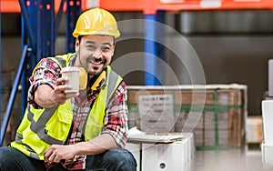 Asian handsome professional male worker smiling, holding cup of coffee, taking rest after work in storage, warehouse or factory