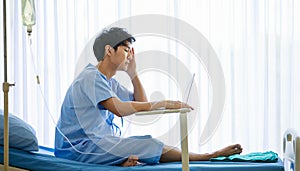 Asian handsome stress male patient wearing blue uniform, getting sick or illness, busy working, using laptop, sitting on bed in