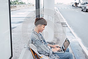 Asian hansome guy is working on a portable computer connected to public wi-fi while sitting on the chair at the airport bus stop