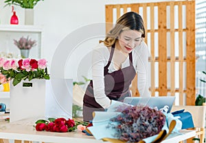 Asian happy cheerful female florist designer shop owner checking smelling quality of red roses blooming in store while using