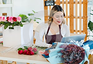 Asian happy cheerful female florist designer shop owner checking smelling quality of red roses blooming in store while using