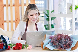 Asian happy cheerful female florist designer shop owner checking smelling quality of red roses blooming in store while using