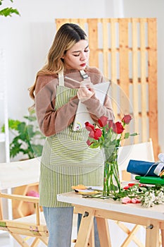 Asian happy cheerful female florist flower shop owner holding pink roses bouquet decorating in white ceramic vase for customer