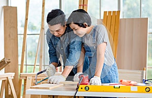 Asian happy cheerful male dad son carpenter woodworker colleague in jeans outfit with safety gloves helping using hand saw cutting