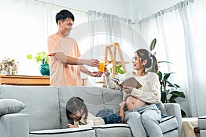 Asian happy family, pregnant mother sits reading book on sofa, with lovely daughter lying next to mobile phone and grabbed mother