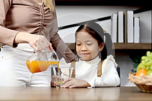 Asian happy family spending time together, mother pouring fresh orange juice from jug for her little cute daughter sitting in